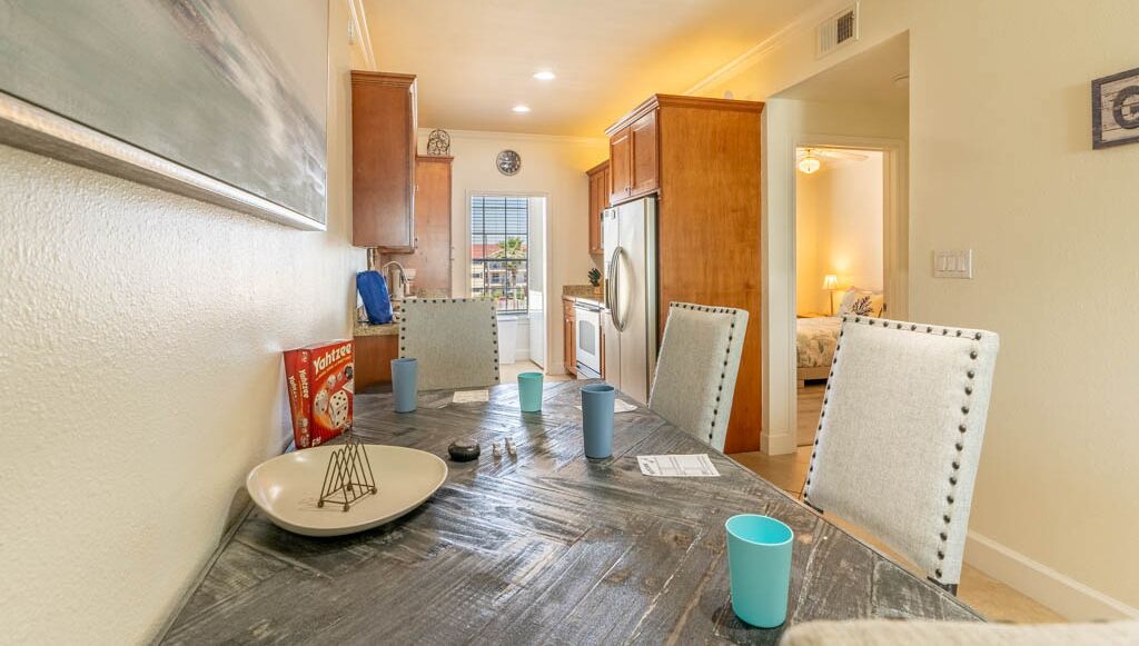 A dining area with a wooden table, chairs, and a plate. There is a refrigerator and a window in the background.
