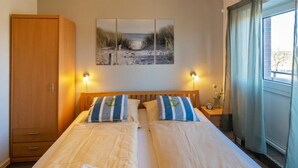 Ein Doppelbett mit zwei Kissen, ein Holzschrank, ein an der Wand befestigtes Kunstwerk und ein Fenster mit Vorhängen.