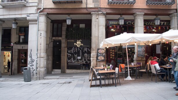 Un café en plein air avec des tables et des chaises, une poubelle et un bâtiment sur lequel on peut lire l’inscription « TURFUTO ».
