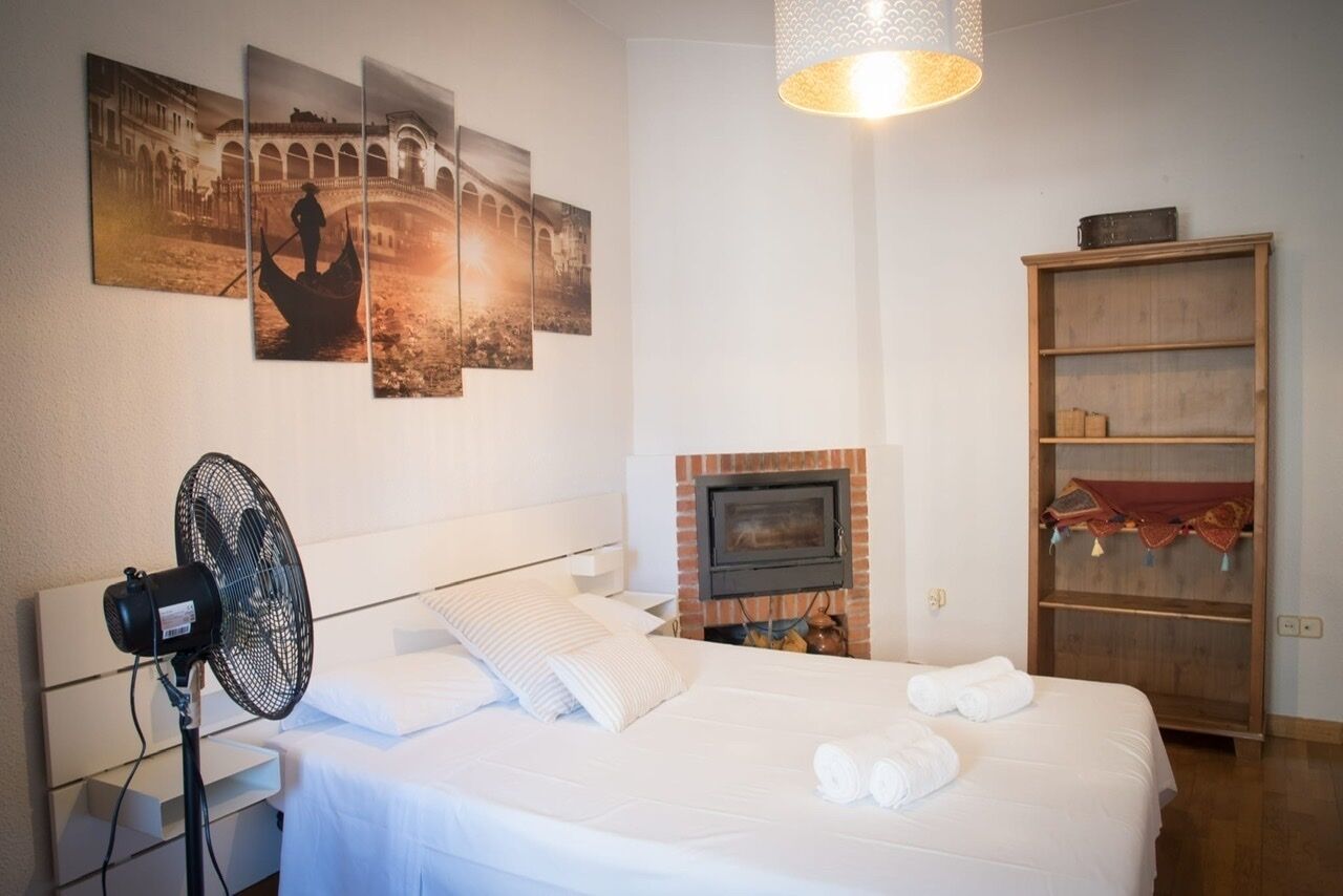 A hotel room with a bed, a standing fan, a fireplace, and wall art depicting a historical scene.