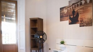 Un lit bien fait, avec des serviettes pliées, un ventilateur sur pied et une étagère murale agrémentée d’une plante.