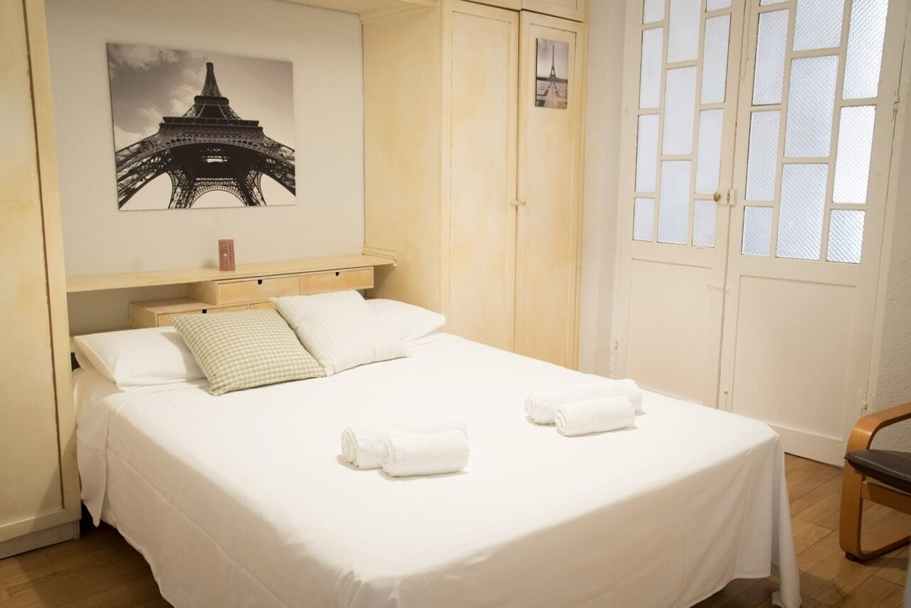 A neatly made bed with white linens and two rolled towels, a wooden headboard, a wall-mounted picture of the Eiffel Tower, and a window with white shutters.