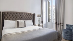 Un dormitorio con cabecera capitonada, cama con ropa de cama blanca y gris, mesita de noche con lámpara, y vistas al exterior a través de una ventana con cortinas.