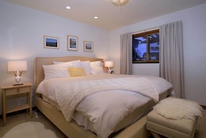 Una camera da letto con un letto grande, due comodini con lampade, una finestra con tende e tre quadri appesi al muro.