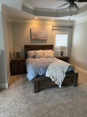 Une chambre à coucher avec un lit, des tables de chevet, un ventilateur de plafond, une fenêtre avec des stores et un tableau au mur.