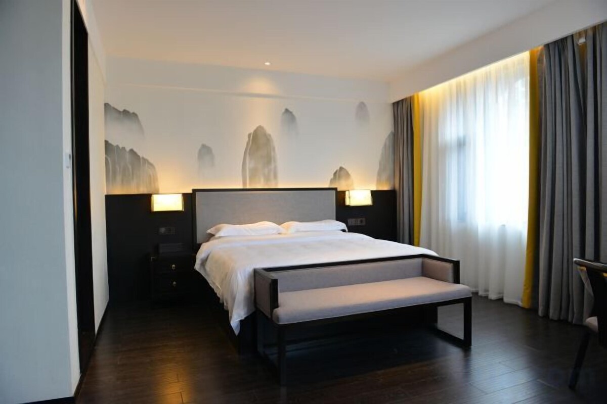 A hotel room with a large bed, bedside lamps, a bench, and a view of the outdoors.