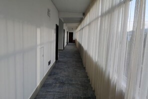 A hallway with a carpeted floor, white walls, and a window with sheer curtains. - Longling Hotel (Baoshan)