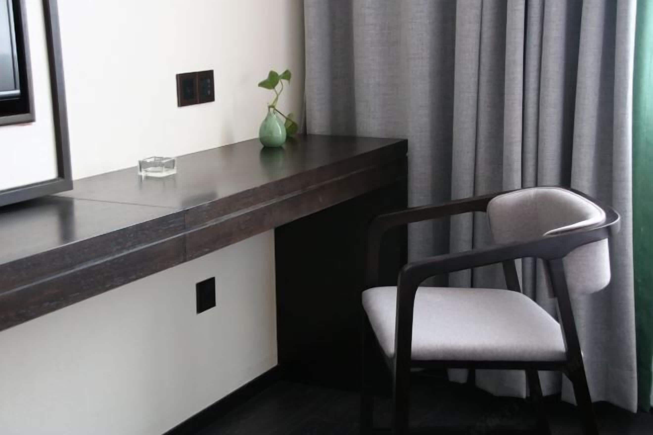 Een moderne hotelkamer met een donker houten bureau, een stoel, een kleine vaas met een plant en een raam met gordijnen.