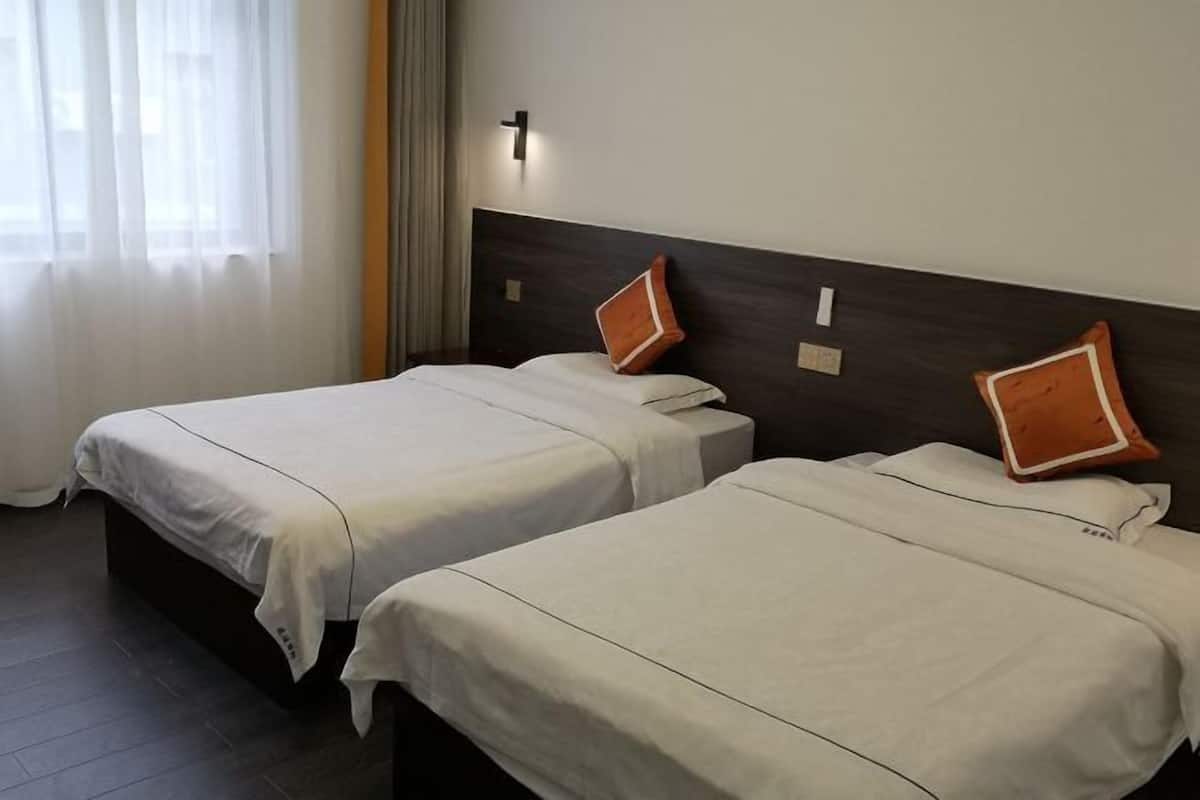 To enkeltsenge med hvide sengetæpper og orange puder i et hotelværelse med en mørk sengegavl, et vindue med lette gardiner og en vægmonteret lampe.