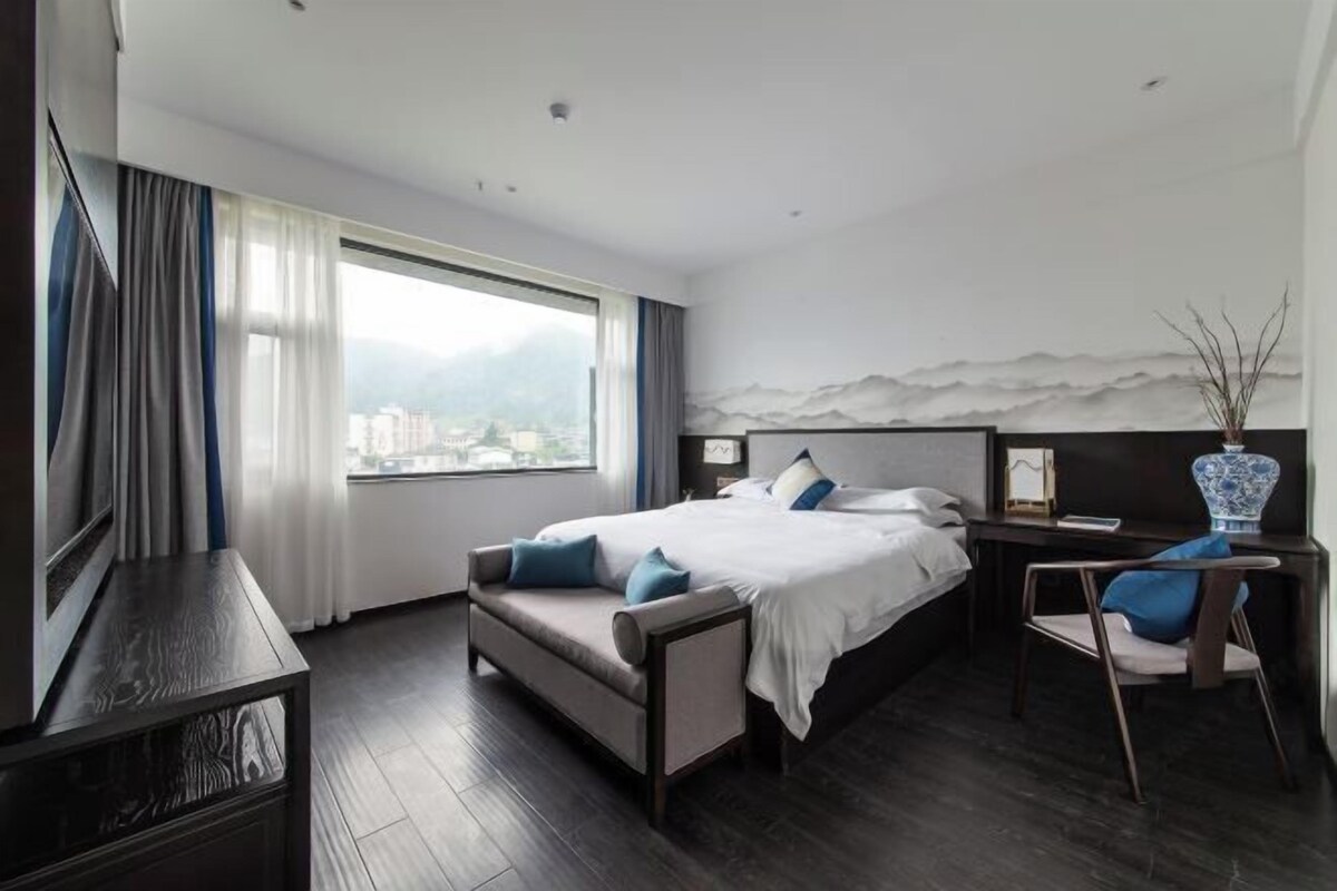 A modern hotel room with a large bed, a desk, and a view of a cityscape through the window.