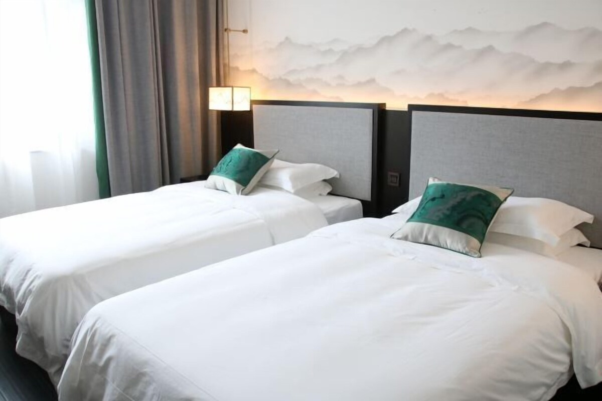 Habitación de hotel con dos camas, cada una con ropa de cama blanca y almohadas verdes. Una lámpara de pared se encuentra a la izquierda de las camas.