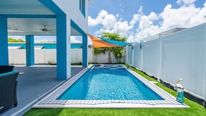 Una piscina con borde de azulejos azules, rodeada por una valla blanca y un área de patio techada con velas de sombra azules y anaranjadas.