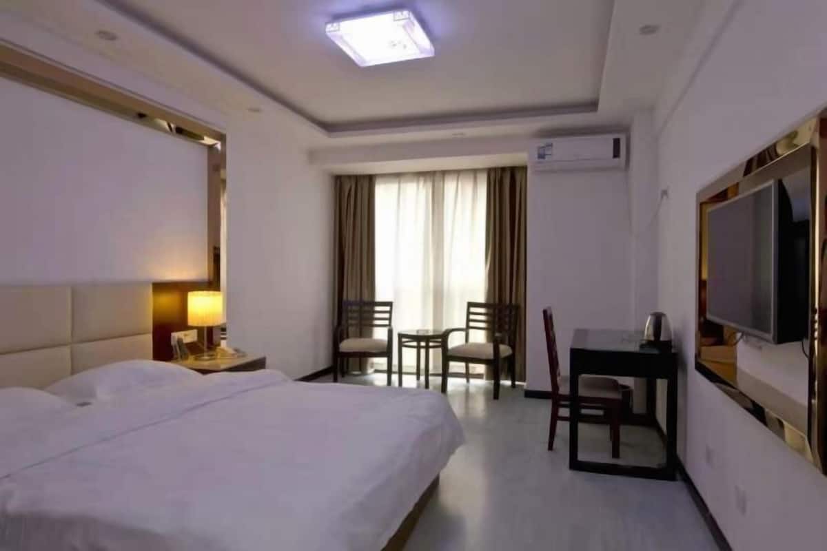 Quarto de hotel com uma cama grande, uma escrivaninha, duas cadeiras, uma televisão e uma janela com cortinas.
