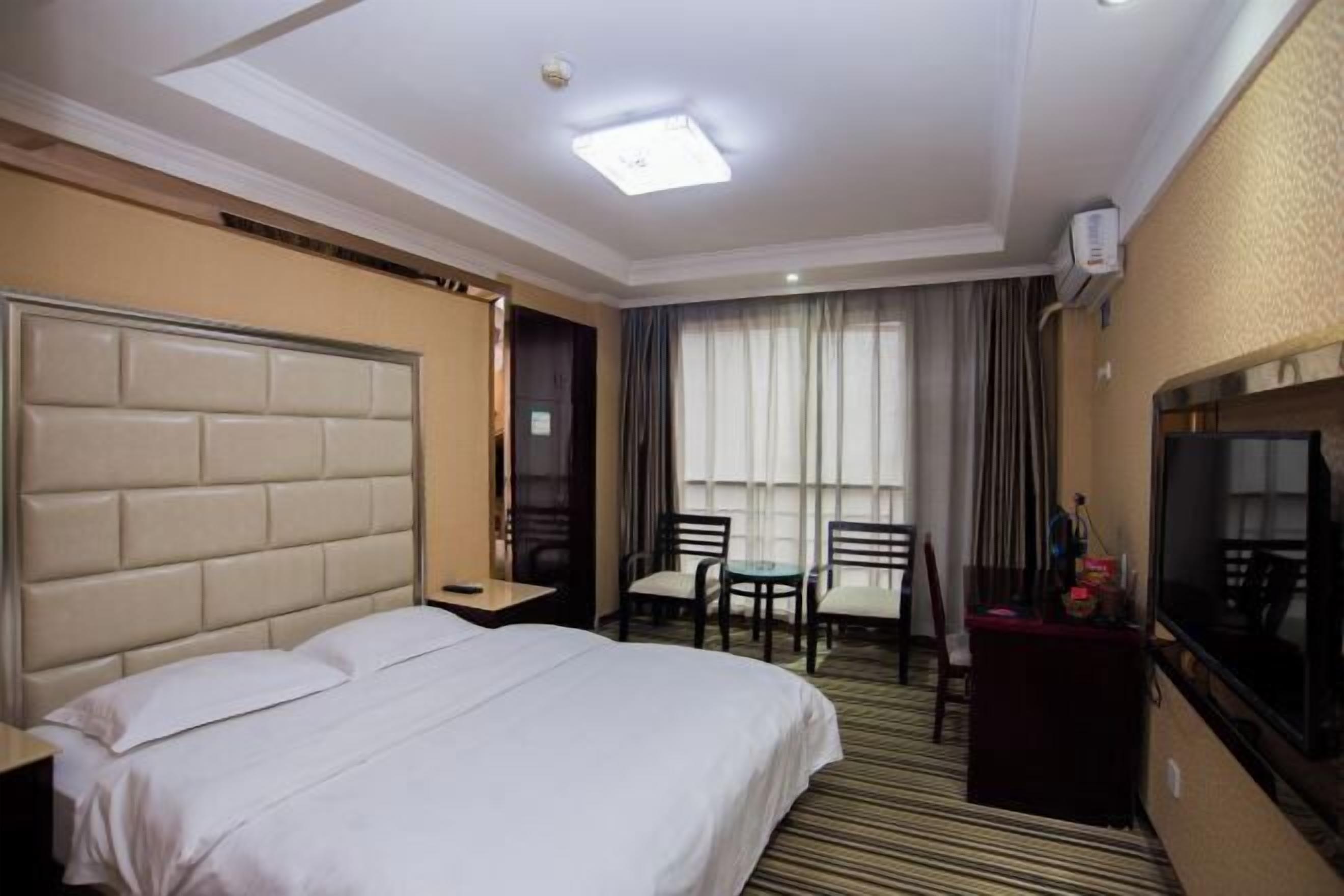 A hotel room with a large bed, a desk, two chairs, a TV, and a window with curtains.