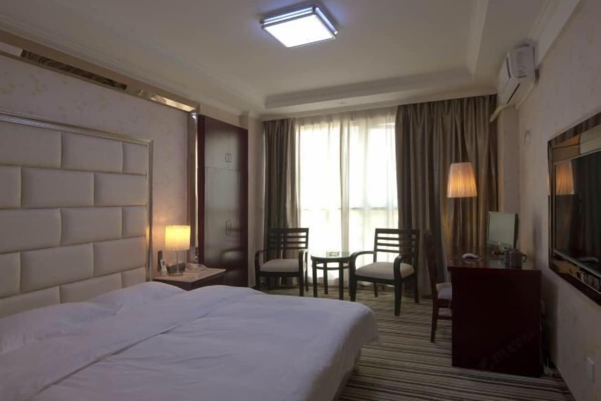 A hotel room with a large bed, a desk, two chairs, a TV, and a window with curtains.