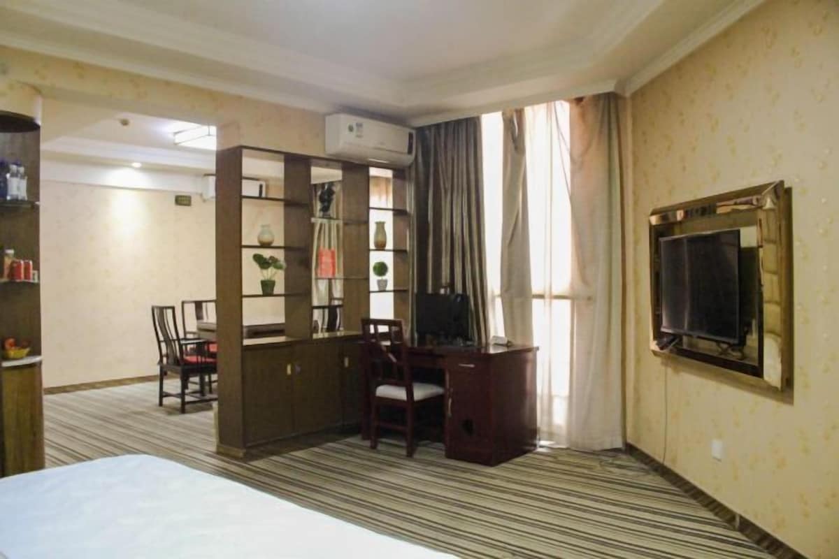 A hotel room with a bed, a desk, a chair, a bookshelf, an air conditioning unit, and a television.