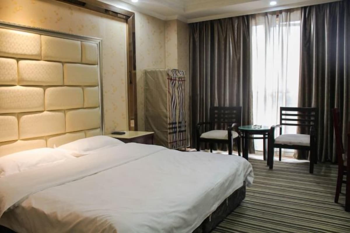 A hotel room with a large bed, a headboard with a textured design, a small table with two chairs, and a view of the outdoors through curtains.