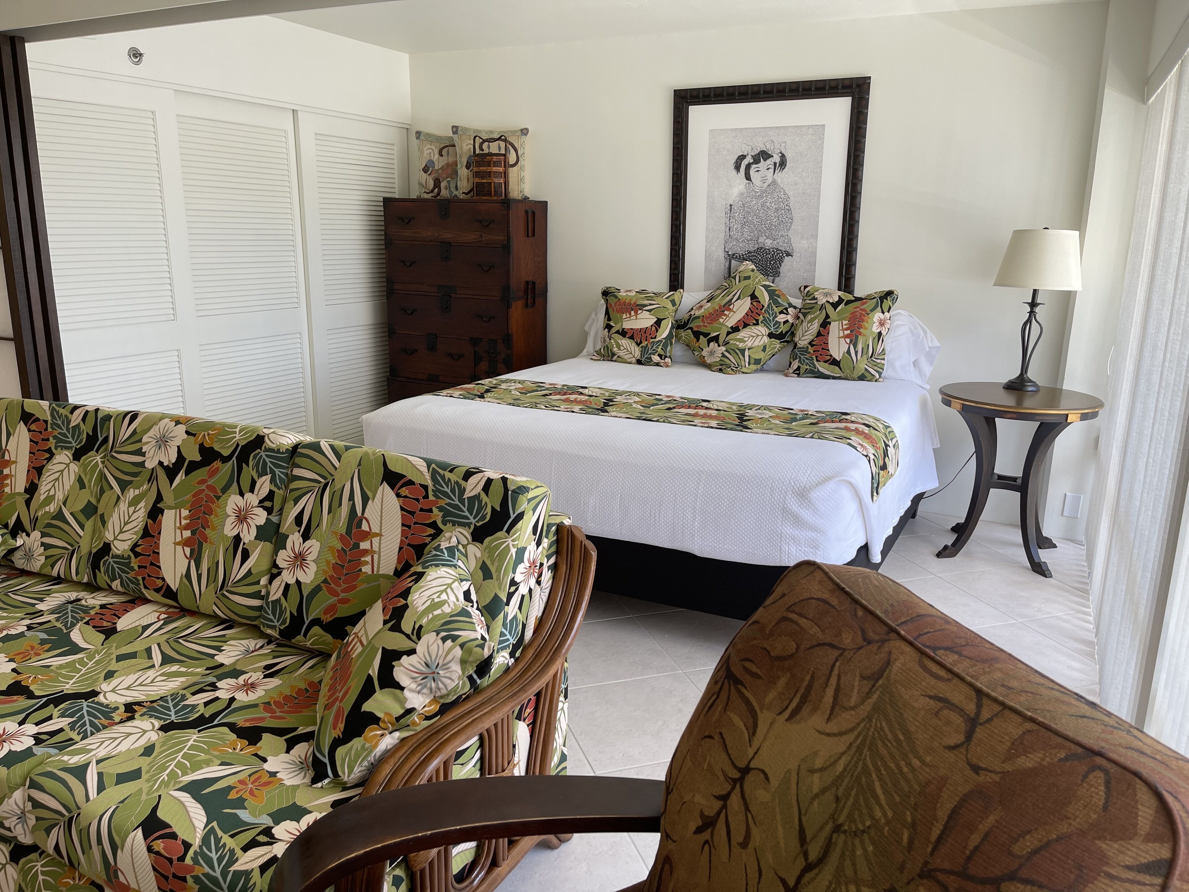 A bedroom with a bed, a floral sofa, a wooden cabinet, a framed picture on the wall, and a small table with a lamp.