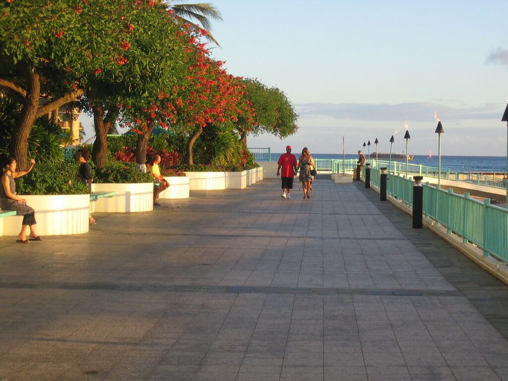 A paved walkway with benches and trees, leading to a waterfront.