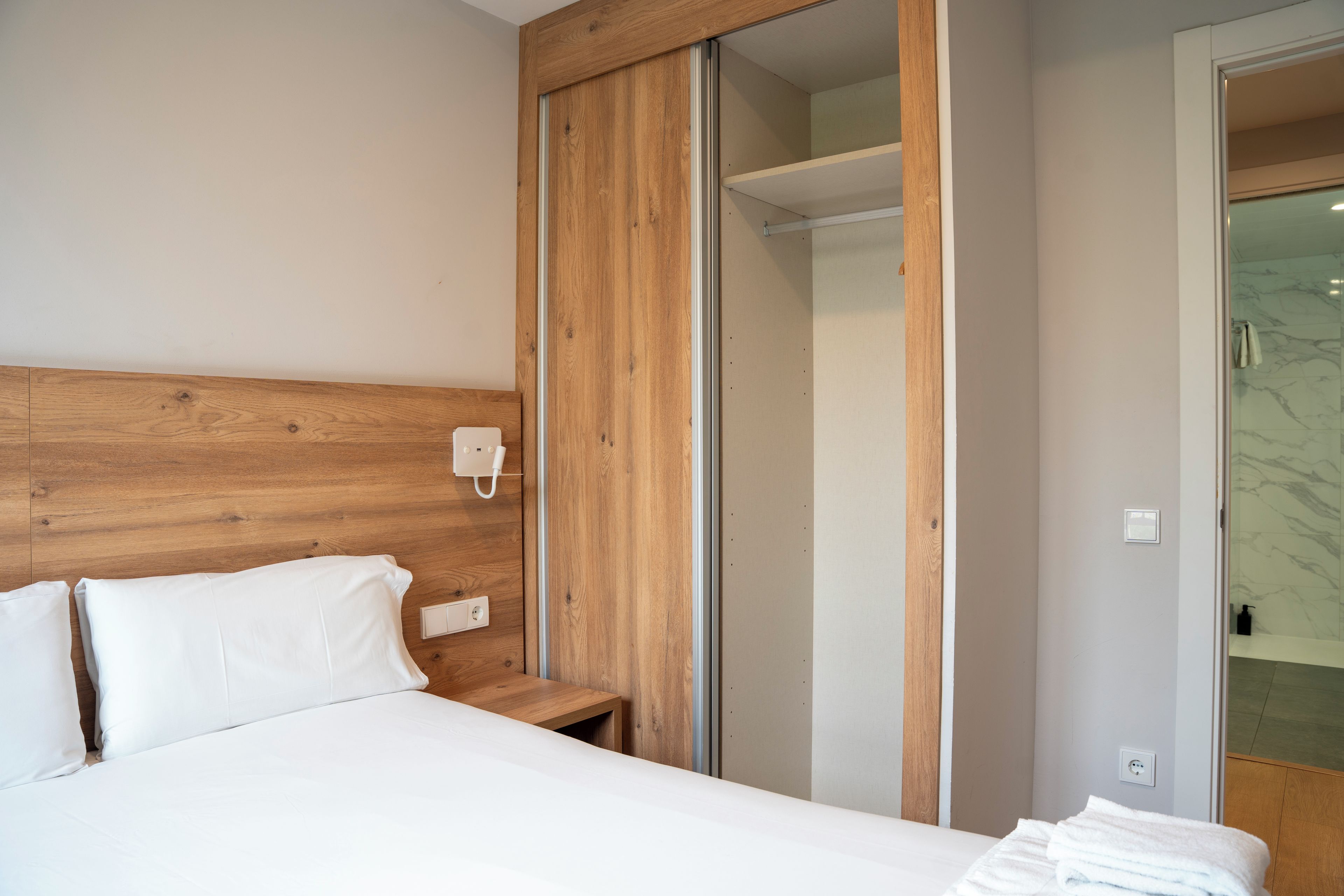 A bedroom with a wooden headboard, a bed with white bedding, a bedside table, and a mirror.