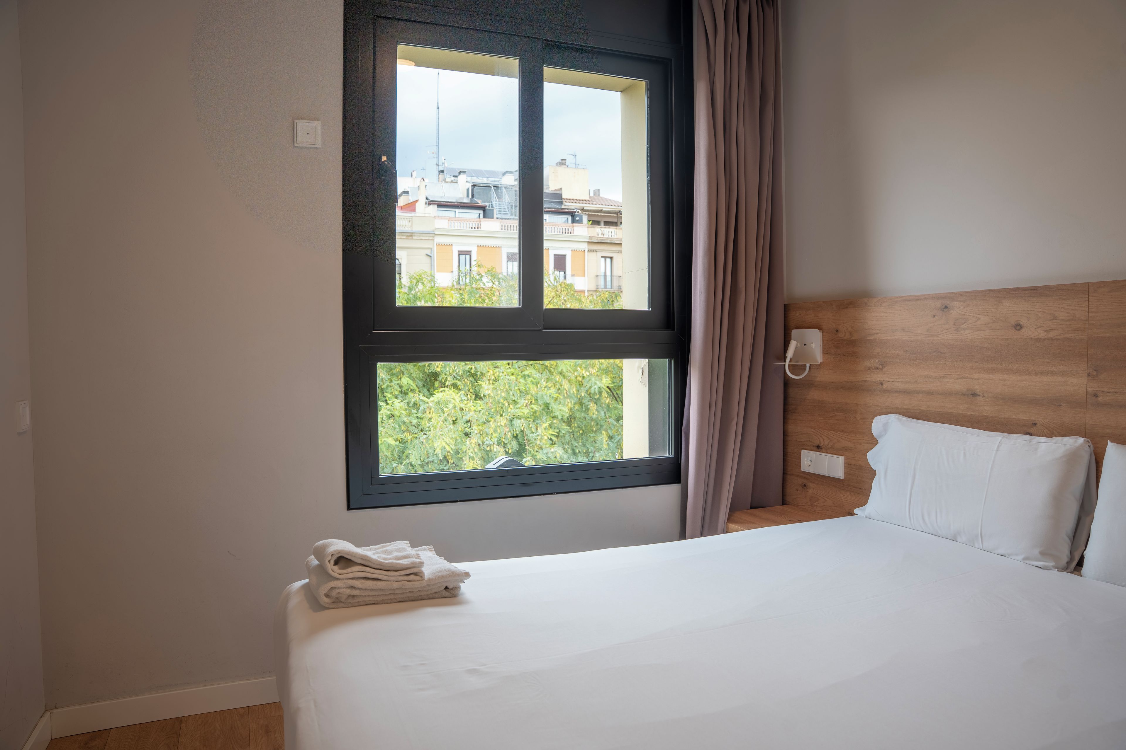 Een hotelkamer met een bed, een raam met gordijnen, uitzicht op gebouwen en een houten hoofdeinde.
