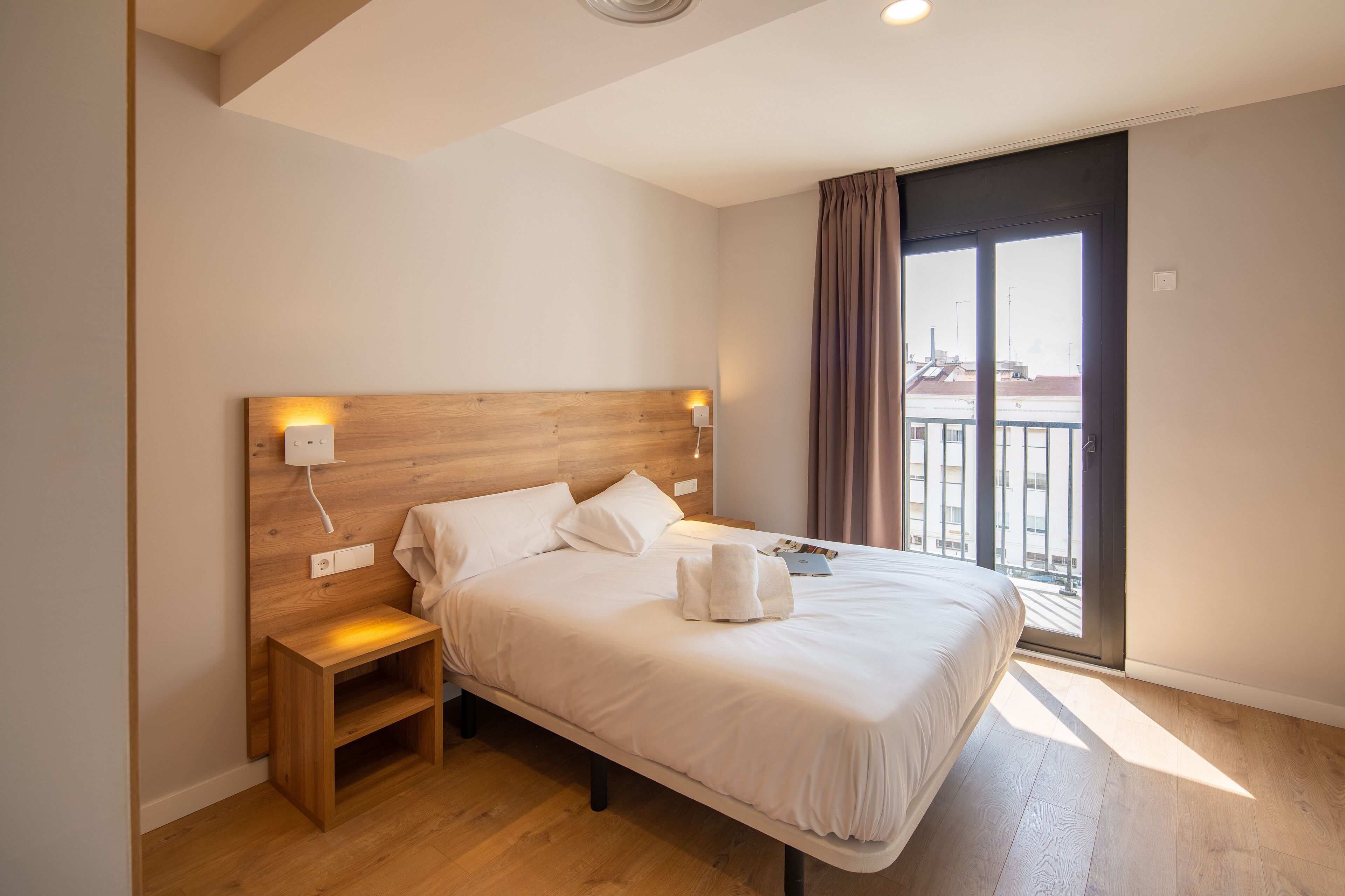 Ett modernt hotellrum med en sänggavel i trä, ett nattduksbord, en stor säng med vita sängkläder och utsikt över staden genom ett stort fönster.