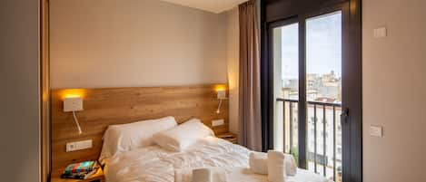 A hotel room with a wooden headboard, a large bed with white linens, two bedside tables with lamps, and a view of the city through a balcony door.
