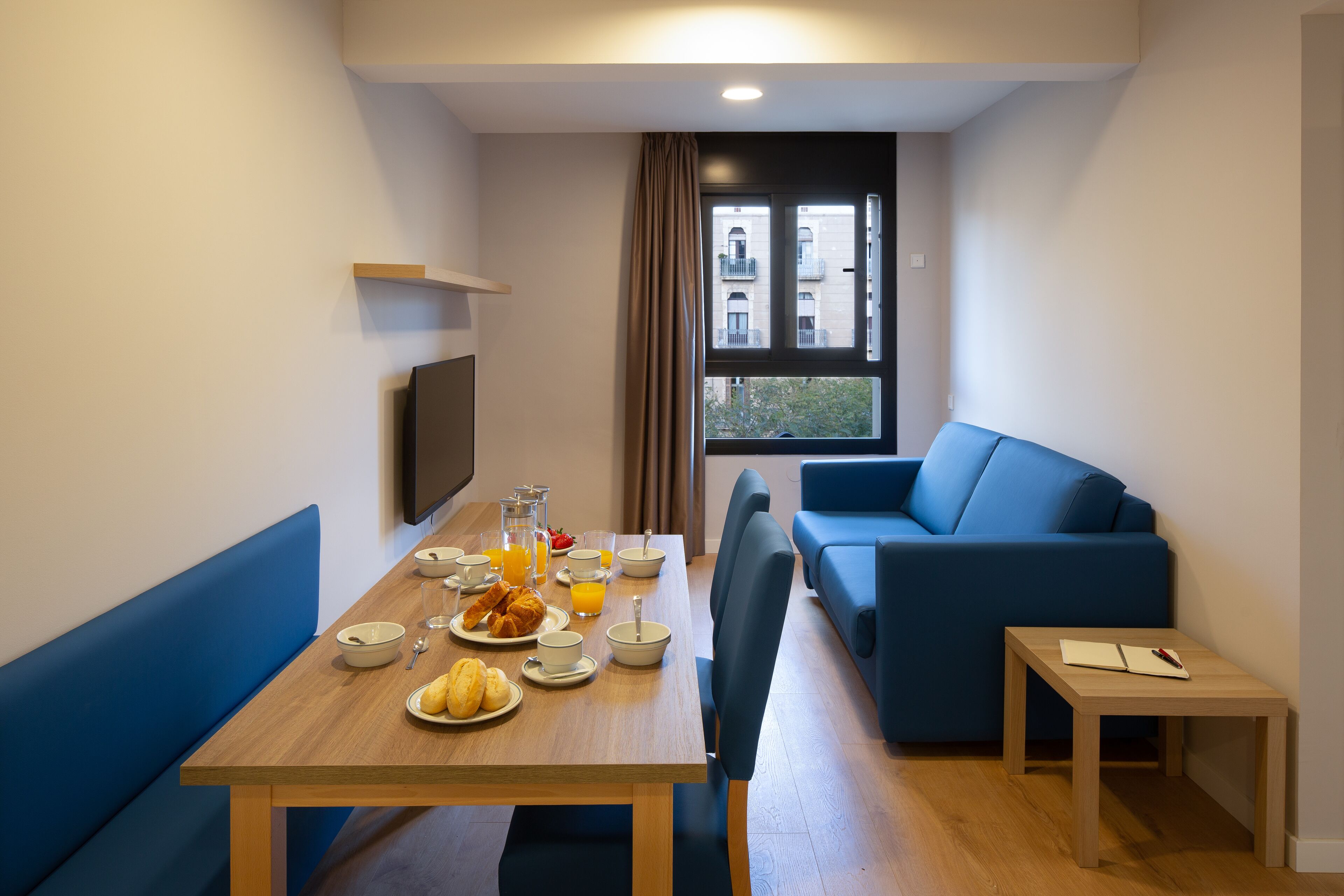 A modern room with a dining table set for breakfast, a blue sofa, and a small wooden table with a book and pen.