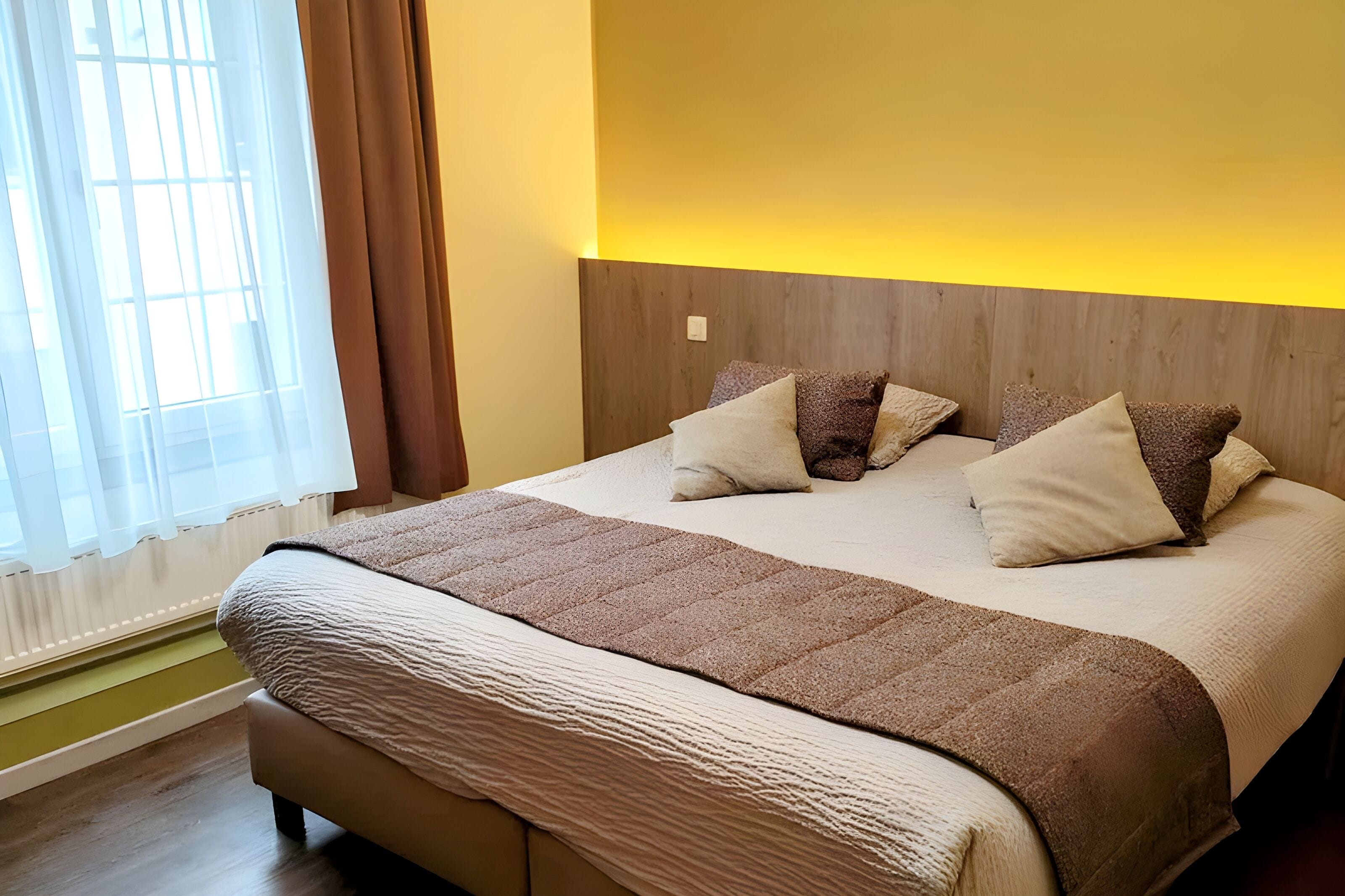 Una cama bien hecha con una manta y almohadas marrones, un cabecero de madera y una ventana con cortinas traslúcidas.