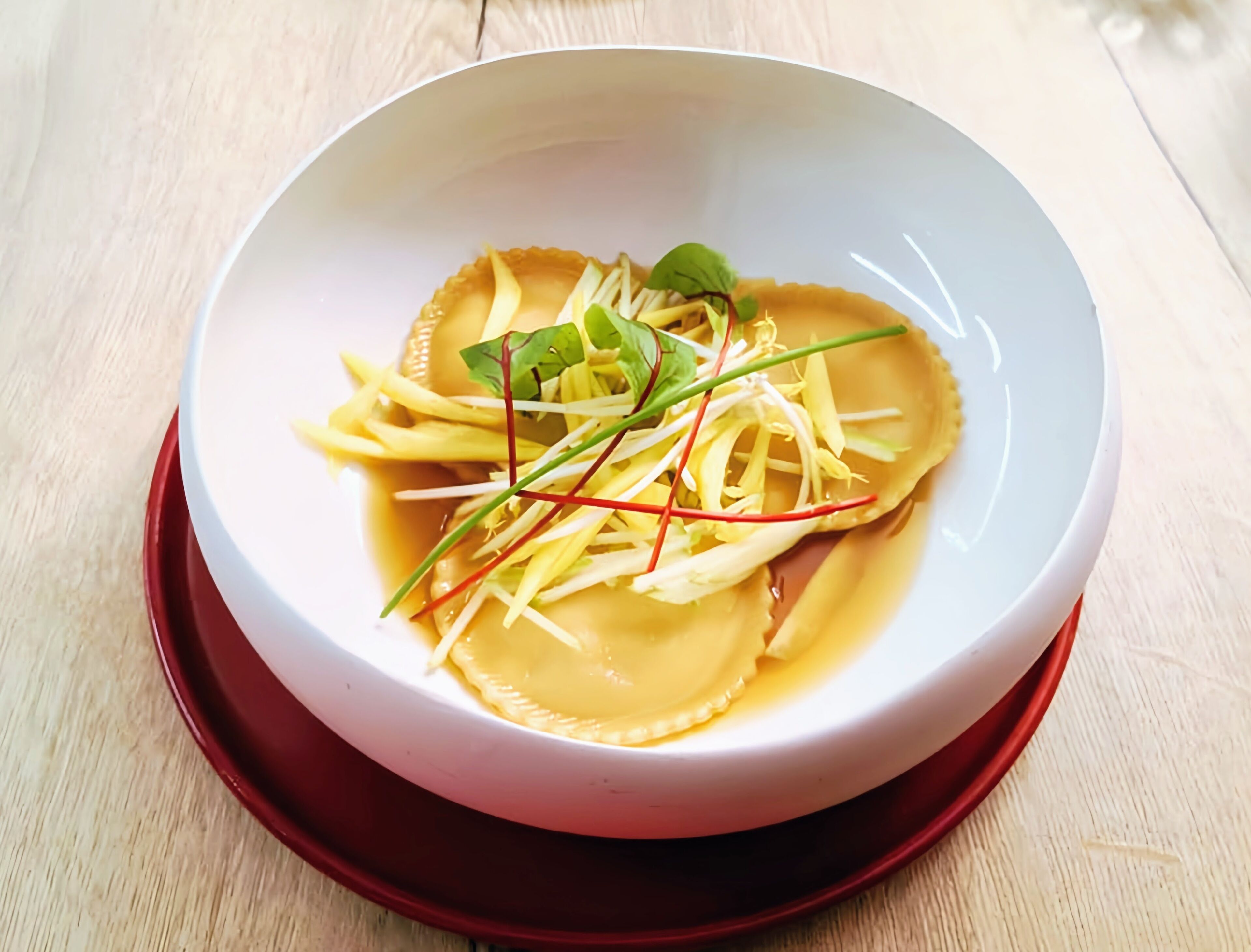 A bowl of pasta with a clear broth and garnishes on a wooden table.