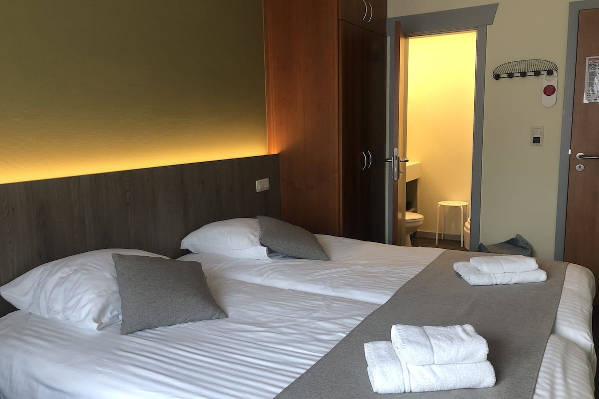 A hotel room with two beds, a wooden headboard, a towel rack, and a door leading to a bathroom.