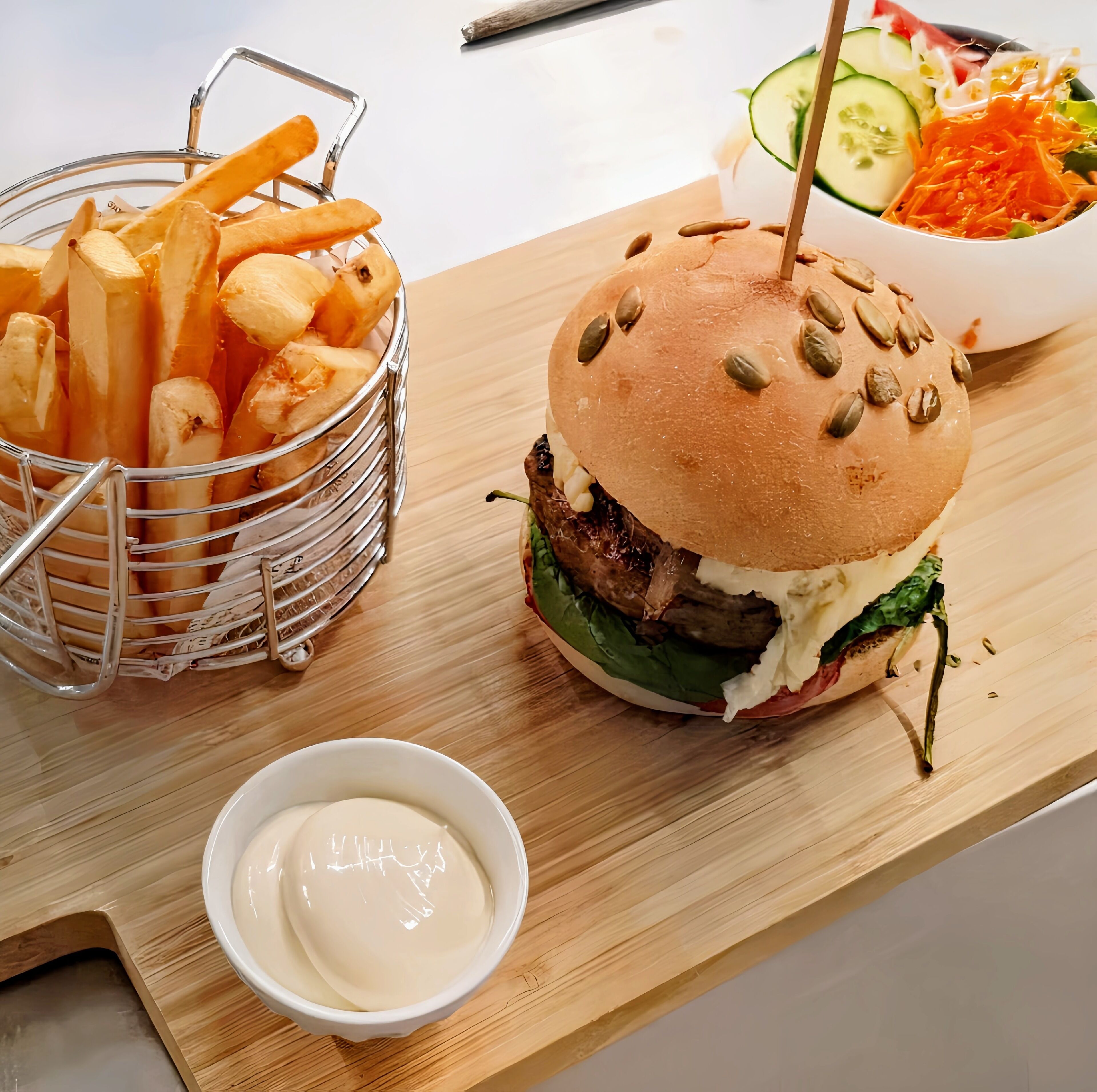 Een sandwich met broodje, vlees en groenten, een saladekom, een mand met frieten en een klein schaaltje saus op een houten plank.