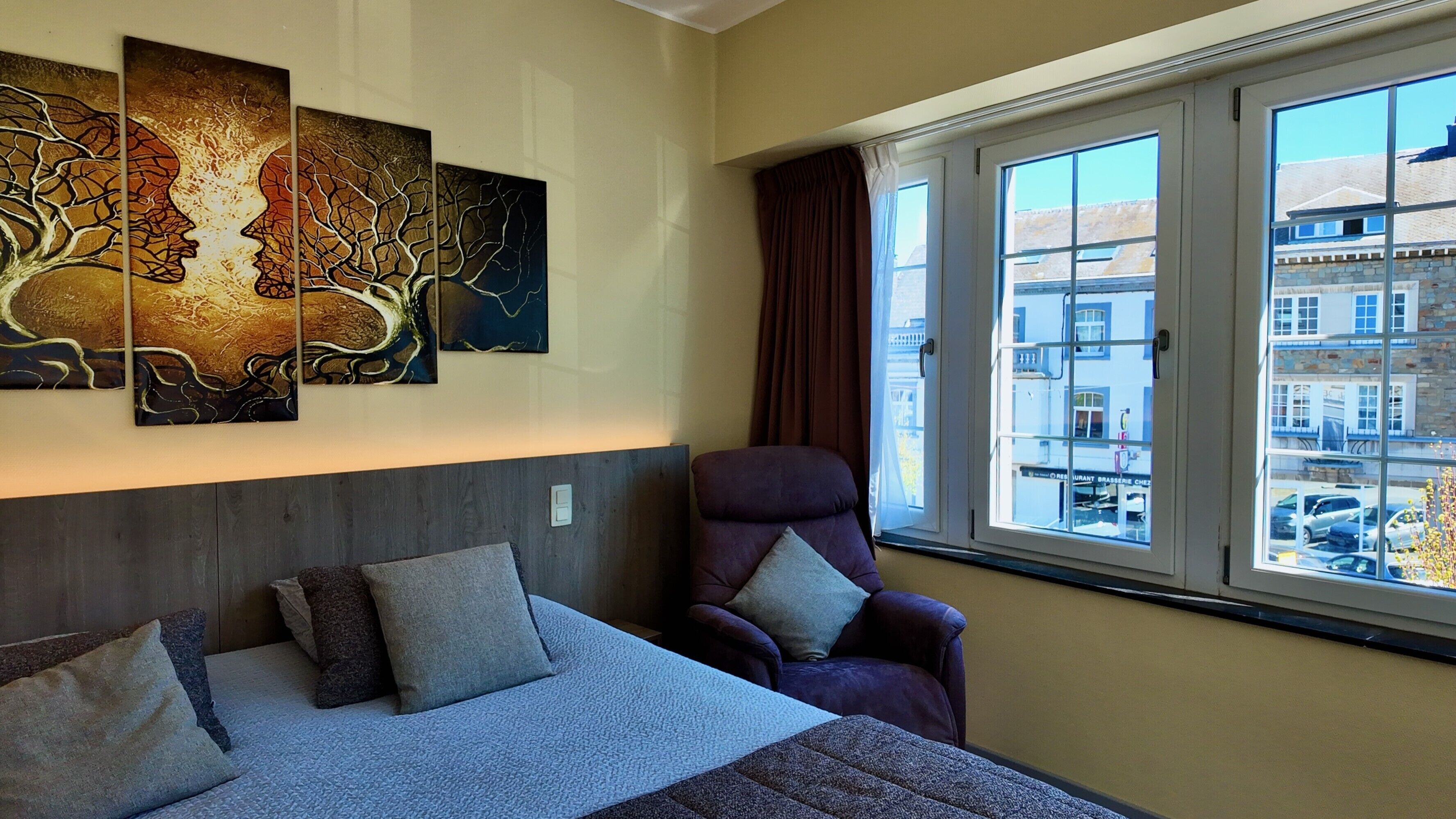 A hotel room with a bed, a chair, a window with a view of buildings, and three artworks on the wall.