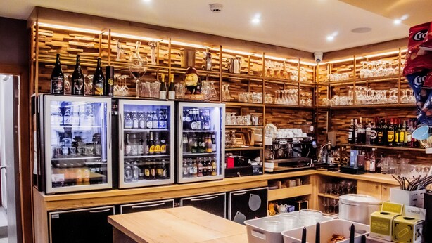 Una zona de bar con refrigeradores llenos de botellas, vasos y un estante de madera con más botellas y copas.