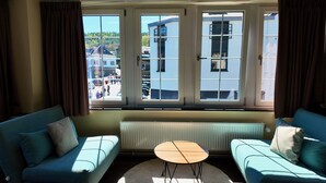Una habitación con dos sofás azules, una mesita redonda y vistas a un edificio a través de amplios ventanales con cortinas.