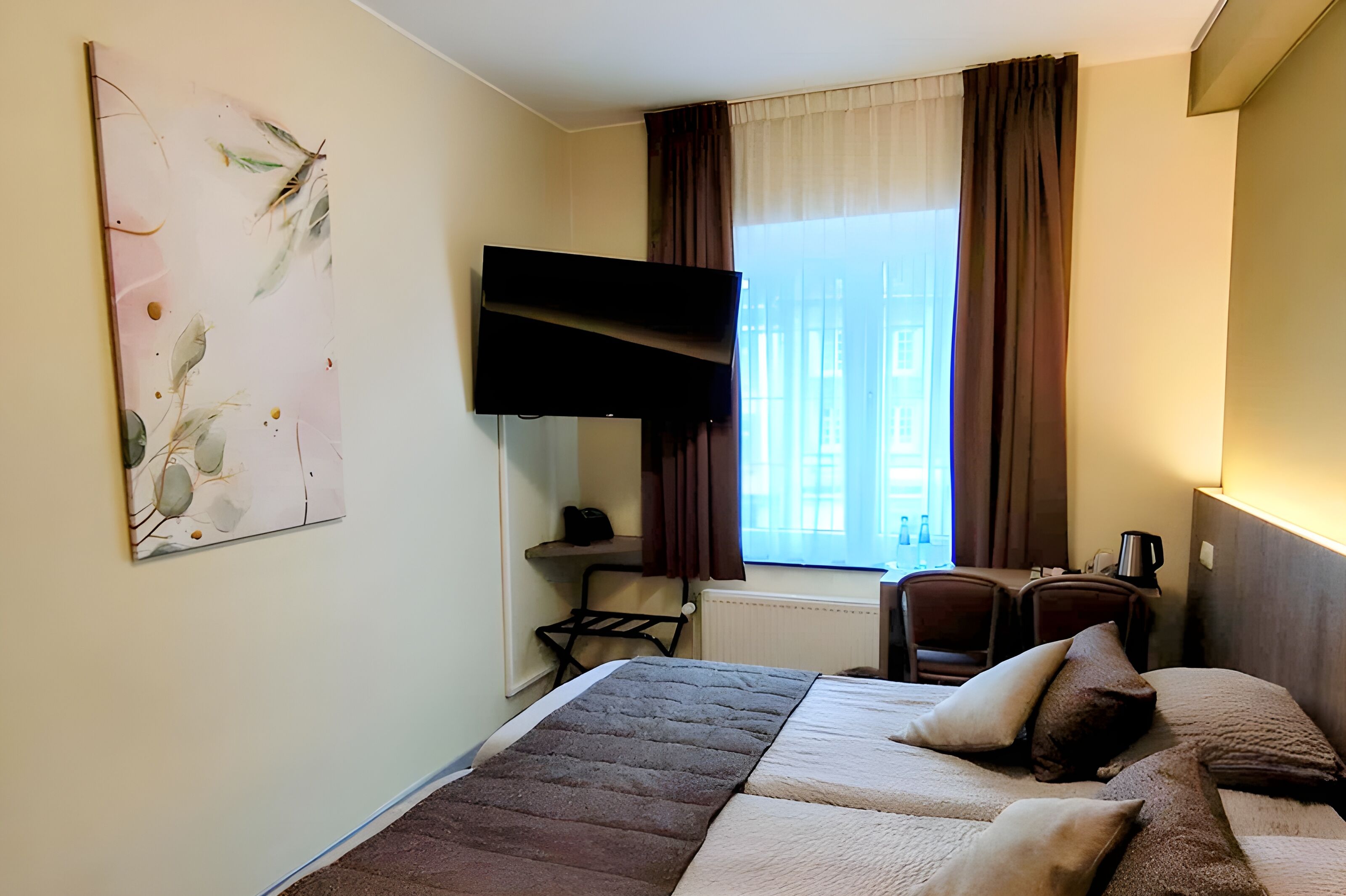 A hotel room with a bed, a desk with a chair, a TV mounted on the wall, and a window with curtains.