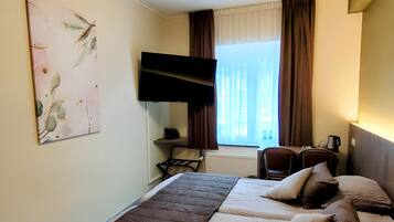A hotel room with a bed, a desk with a chair, a TV mounted on the wall, and a window with curtains.