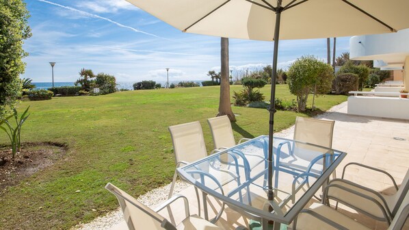 Une table en verre entourée de chaises, placée sous un grand parasol, avec vue sur une pelouse arborée et la mer.
