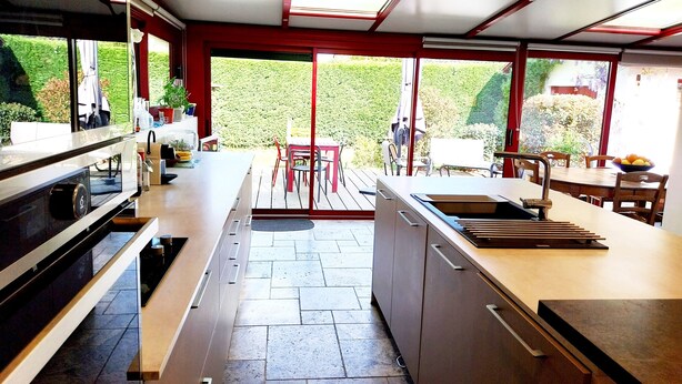 Una cocina moderna con una isla central, un comedor con mesa y sillas, y vistas a un jardín a través de amplios ventanales.