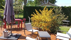 Una terraza de madera con mobiliario exterior, incluyendo sillas y una mesa, rodeada de follaje y un seto.