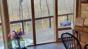 A wooden cabin room with a large glass door leading to a deck. A table with a glass top and a flower vase is in the foreground.