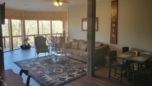 A spacious living room with a large window, a ceiling fan, a wooden floor, a patterned rug, and a dining area with a table and chairs.