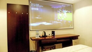 A hotel room with a bed, a desk with a laptop, a projector screen displaying a sports game, and a wooden wardrobe.