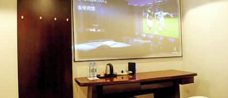 Ein Hotelzimmer mit Bett, Schreibtisch mit Laptop, einer Projektionsleinwand, die ein Sportspiel zeigt, und einem Holzschrank.