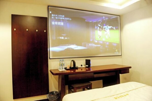 Une chambre d’hôtel équipée d’un lit, d’un bureau avec un ordinateur portable, d’un écran de projection diffusant un événement sportif et d’une armoire en bois.