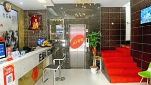 A hotel reception area with a reception desk, a digital screen, a red carpeted staircase, and a television on the wall.