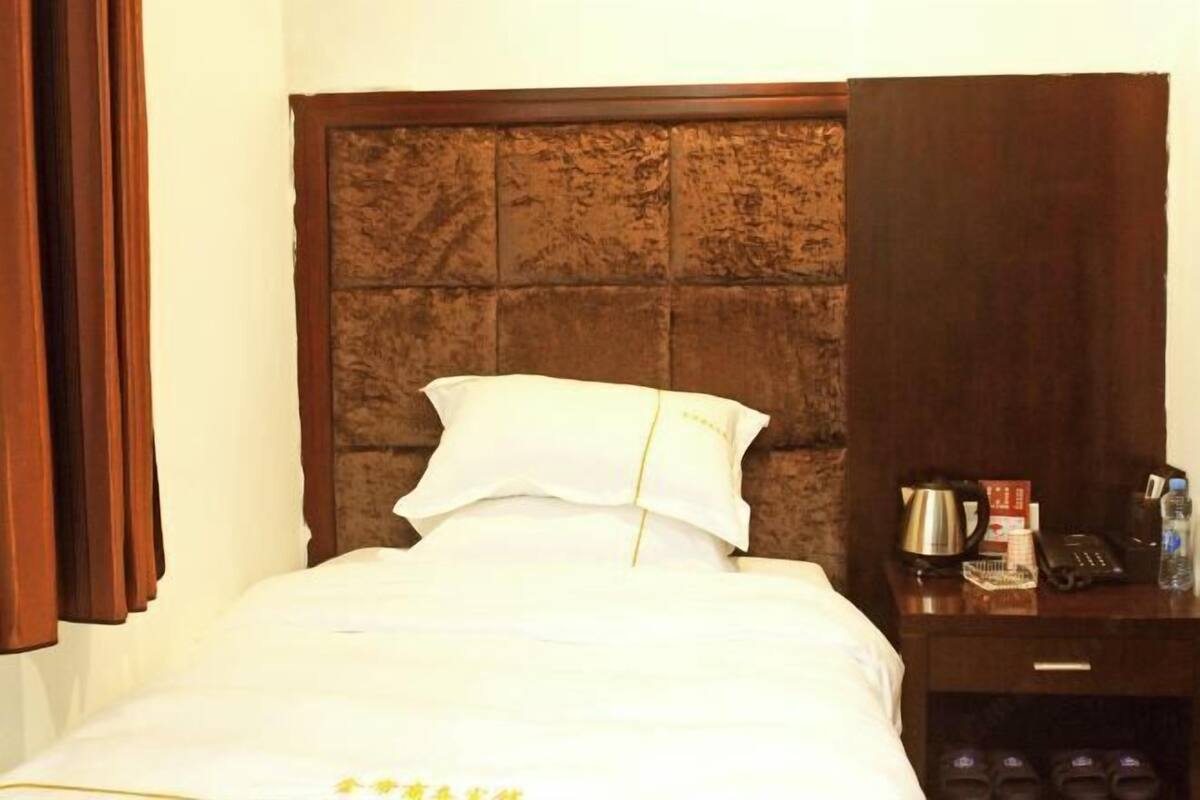 A hotel room with a large bed, a wooden headboard, a nightstand with a kettle, a phone, and a water bottle.