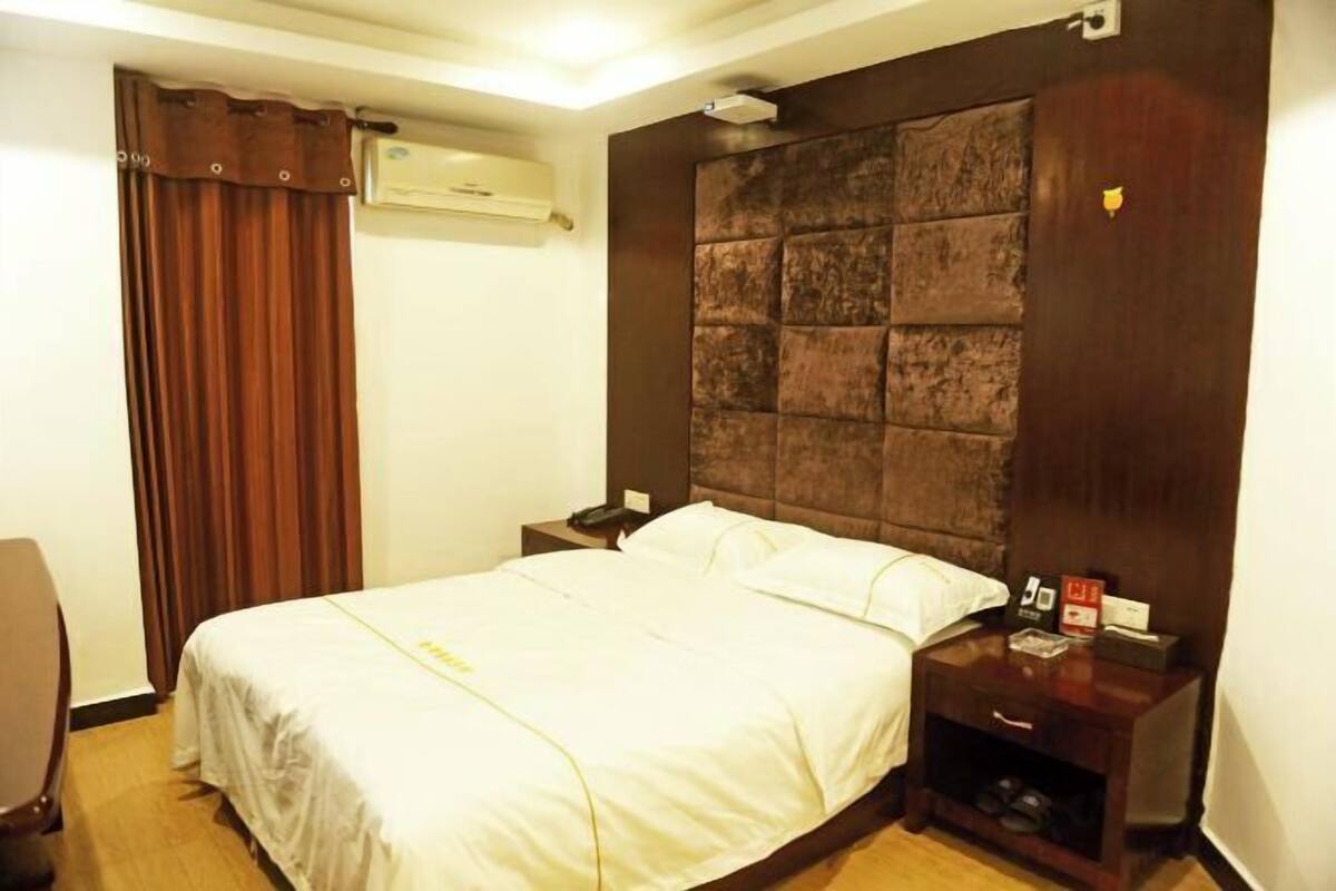 A hotel room with a large bed, a wooden headboard, a bedside table with a phone and a lamp, and a curtain on the window.