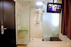 Une chambre d’hôtel comprenant un lit, un téléviseur fixé au mur, un espace douche et un petit tabouret.