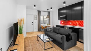 Ein modernes Wohnzimmer mit einem grauen Sofa, einem Glastisch und einem Fernseher. Es gibt eine Küche mit schwarzen Schränken und einer roten Spritzschutzwand.